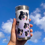 Hand holding a Wrapware tumbler covered in dog portraits against a blue sky.